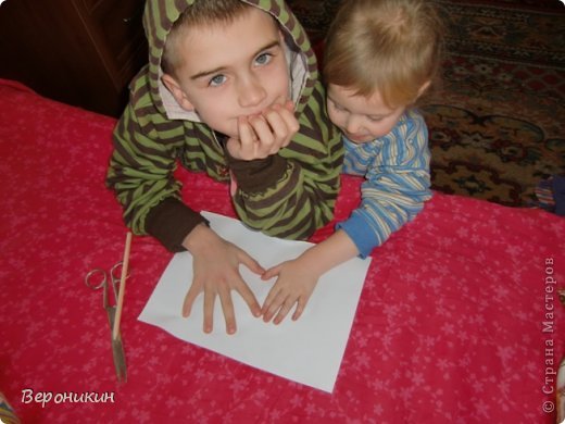 Сначала помогите деткам положить ладошки на лист бумаги так, чтобы между двумя ладошками получалось сердечко. У меня особенно интересно - детки разного возраста. Обведите карандашом. Из картона зеркально вырежьте для открытки две такие детали - на одной нужно прорезать получившееся сердечко, а на второй - оставить. На первой детали внутри сердечко можно украсить бусинами, нанизанными на ниточку. Мне детки сами подавали бусинки, которые им больше нравятся. На вторую деталь открытки на внутреннюю сторону, в том месте, где не вырезано сердечко нужно наклеить нарядный фон - у меня самоклеющаяся плёнка. И соединить обе детали открытки клеем.  (фото 2)