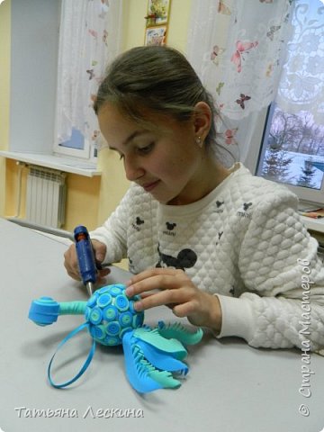 6. Для шеи понадобилось: проволока нужной длины, отрезок фоамирана бирюзового цвета примерно 10*12 см. Проволоку приклеили к фоамирану таким образом, чтобы её было немного видно с обеих сторон, скрутили прямоугольный отрезок в тугой цилиндр, склеили клеевым пистолетом. Приклеили к голове(отрезок проволоки вошёл внутрь головы) Шею с головой приклеили к тельцу-шарику, при этом отрезок торчавшей проволоки вошёл в пенопласт. (фото 10)